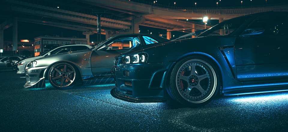 Nissan Skyline R34 GT-Rs mit Neon-Unterbodenbeleuchtung aufgereiht bei einem nächtlichen Auto-Treffen an der Daikoku PA unter Autobahnbrücken
