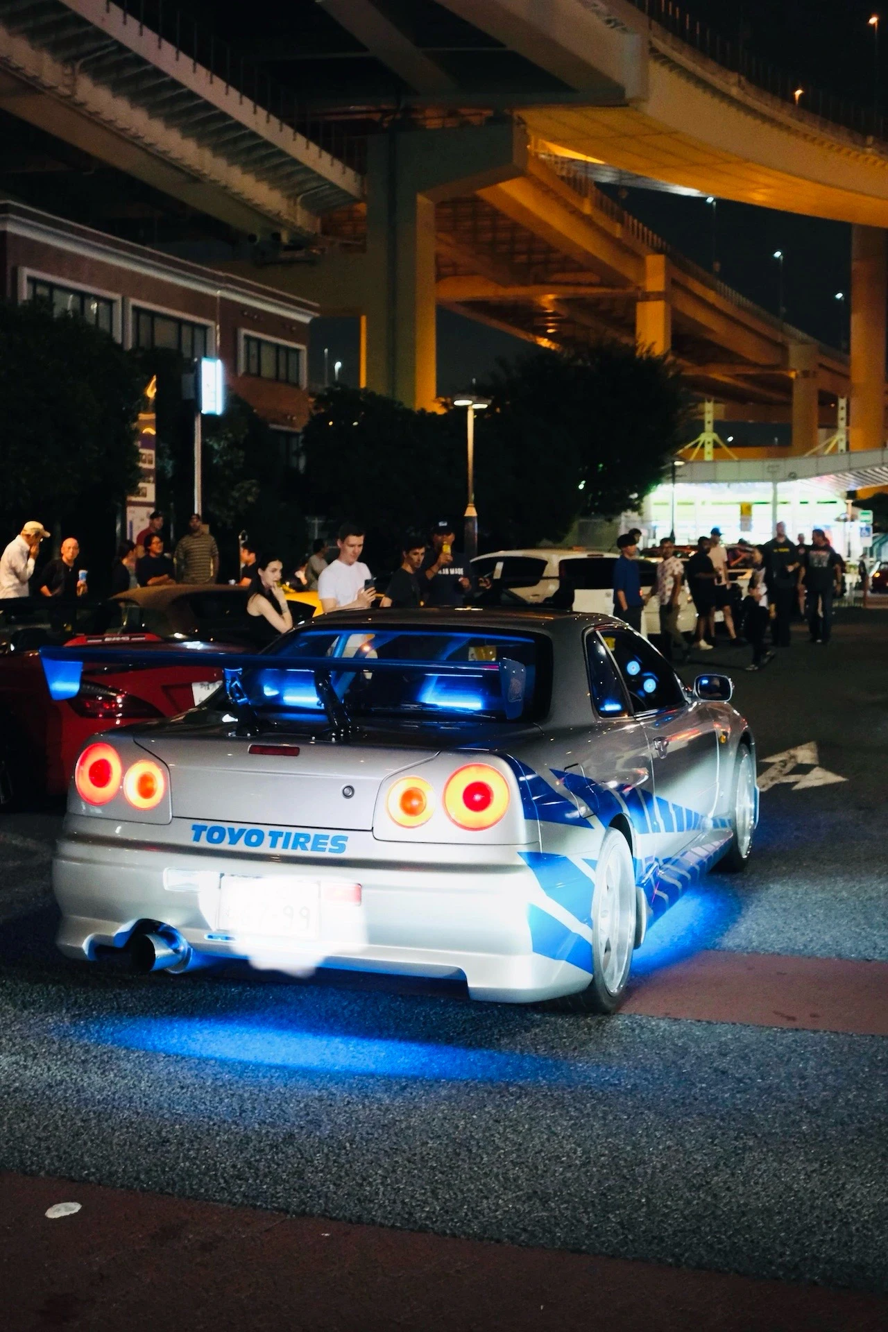 Silberner Nissan Skyline R34 mit blauer Unterbodenbeleuchtung, umringt von Zuschauern beim Auto-Treffen an der Daikoku Parking Area