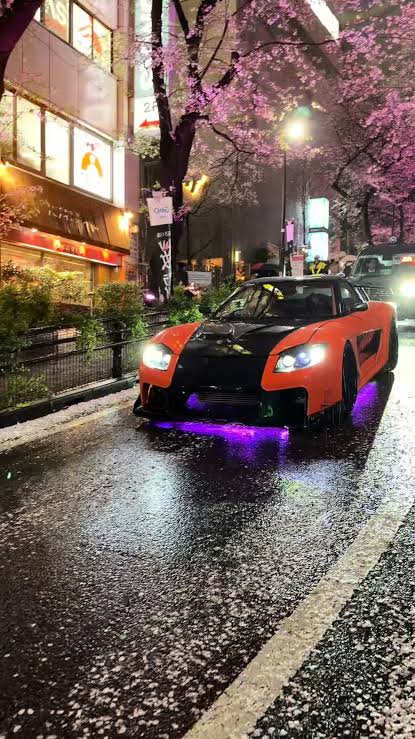 Oranger Veilside RX-7 fährt nachts bei Regen unter Kirschblüten durch eine Straße in Tokio