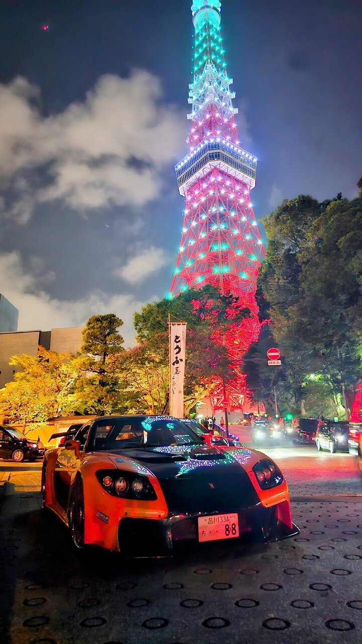 Mit Lichtern dekorierter Veilside RX-7, nachts vor dem beleuchteten Tokyo Tower geparkt